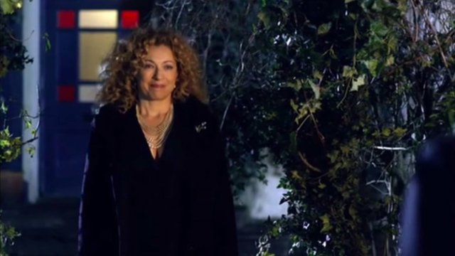 River Song stands outside wearing nice black clothes and pretty jewelry