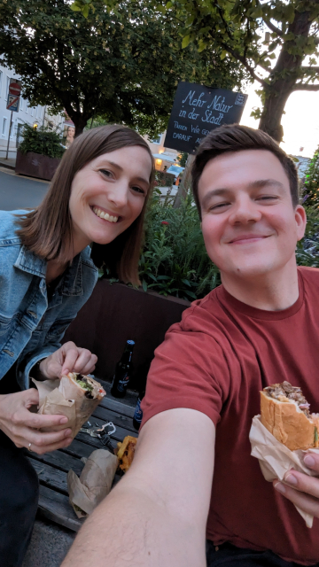 My fiancée Claudi and I sitting on a public bench smiling and eating fast food. Selfie.