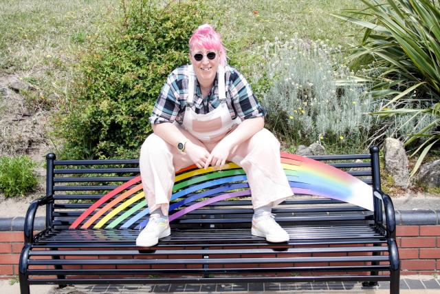 Alexis, a woman with light skin tone and pink hair, wearing sun glasses, a flannel shirt and dungarees, sits on the back of a rainbow bench with her legs wide, her arms leaning on her thighs.
