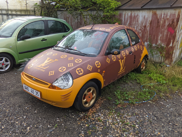 A second generation of the Ford Focus car with a paint job in Louis Vuitton purse design.
It has the typical brown and orange colors with many snowflakes around the car. There is a huge LV logo with smaller czech text saying "Luksusní Vrak".