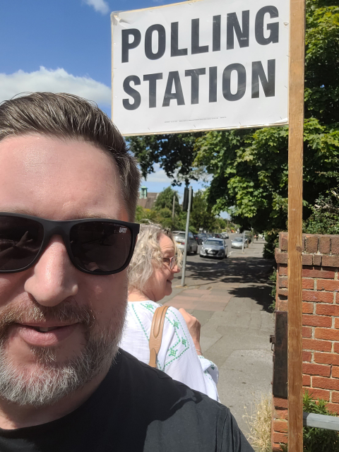 I'm just a man. Standing in front of a camera. Asking you to love me.

Its me standing in front of the polling station sign outside a local school whilst a woman smirks in the background. Probably because I had just said to her in an unbearably cringey way "It's for the 'gram". What a absolute prick. I've got a black tshirt and black sunglasses on. My hair looks like a velcro wig but also simultaneously greasy somehow. I have a beard, which barely conceals some of the less kind contours of my big face. A face that looks like a boxer who's never been in a fight. The face of a kindly sailor who never went to school but knows the mysterious ways of the ocean. Like a man who expected to be famous but never was but still behaves like a celebrity because he doesn't know anything else. Is it a kindly face? It's THE kindly face. If you had a fleet of puppies and needed them looking after, this would be the face you'd choose to protect your puppies. Would you give him a simple task to do, expecting it to be carried out to instructions? Absolutely not. You would expect to ask him to hand you a slice of bread and then turn around to find he'd disappeared, turning up 8 hours later in another town with no trousers on and covered in bees. A kindly idiot. A sort of philosopher poet who never made sense in his lifetime but found centuries later to be the greatest thinker of the age. A street magician with no magic. The plucky but ultimately hapless minor love interest in a marvel film. 