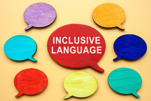 An image features a yellow background with eight colorful wooden speech bubbles arranged in a circular pattern. The speech bubbles are painted in various colors: purple, orange, blue, teal, green, yellow, red, and another red one in the center. The central red speech bubble has the words 'INCLUSIVE LANGUAGE' written in white capital letters. The arrangement and bright colors create a vibrant and engaging visual emphasizing the importance of inclusive language.