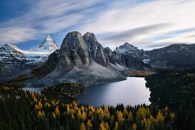 A grand landscape scene of the Assiniboine Mountain range. Three Lakes can be seen in the foreground. The forest is speckled with yellow larche trees.