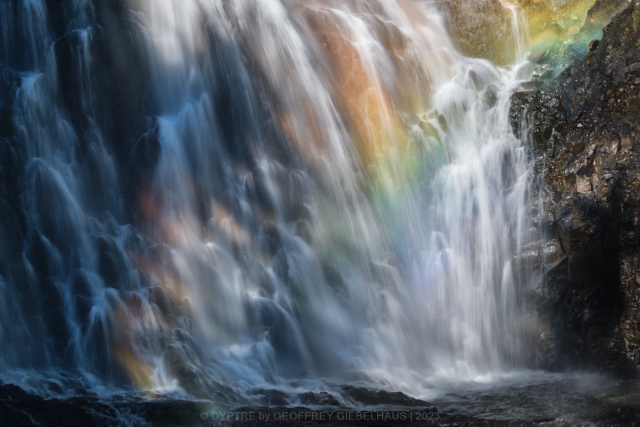 A closeup of a large waterfall. Midday sunlight illuminates the mist creating a rainbow of colour in the scene.