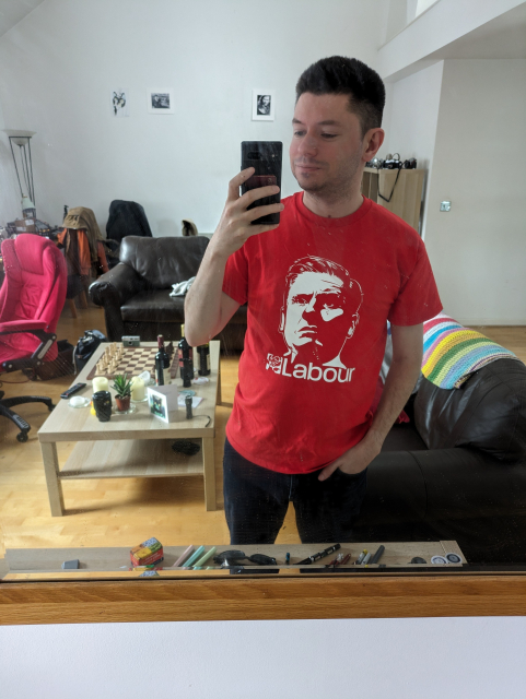 A picture if me in my living room mirror wearing a bright red Labour tshirt with Keir Starmer's face emblazoned on it.  In my room, a coffee table with a chess board, some bottles of wine, and plants is visible.