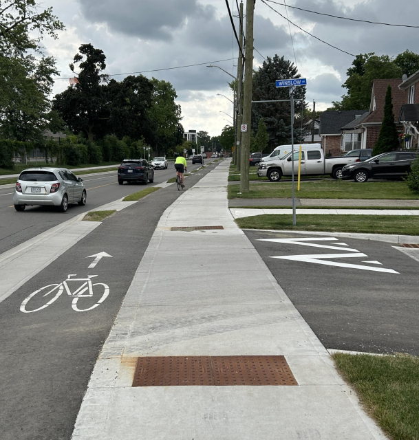 A cyclist rides in a designated bike lane alongside a road with cars. The sky is overcast, and houses with lawns line the street.