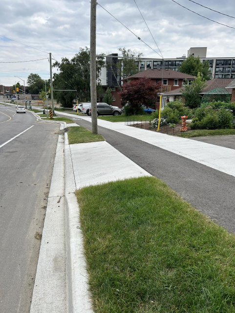 A suburban street with a sidewalk and grassy verge. Residential houses, cars, and utility poles are visible, along with trees and a building in the background under an overcast sky.