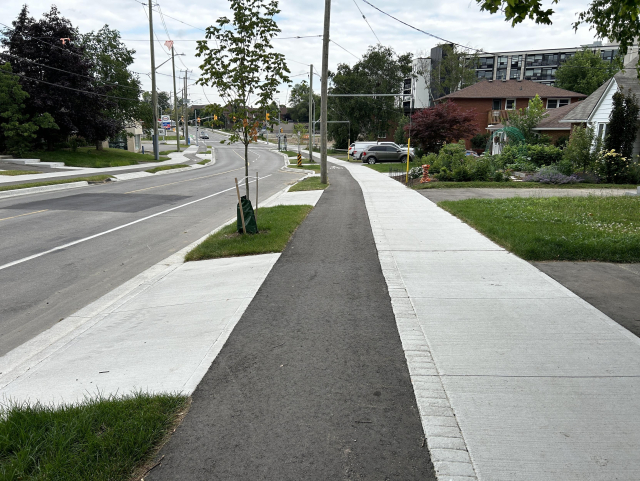 A freshly paved residential street with new sidewalks, young trees planted along the walkway, houses with gardens on the right side, and power lines overhead.