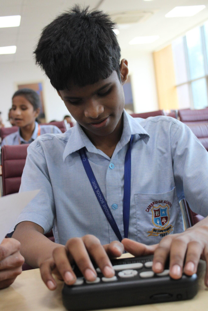 A student from Cambridge School Noida is seated in a classroom, using a braille device. He is wearing a light blue uniform shirt with the school emblem and a blue lanyard. He is focused on the braille device in front of him, pressing the buttons with both hands. Another student is visible in the background.