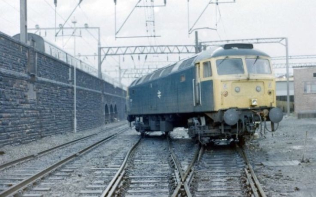 Rail blue BR Class 47 where the points must've moved under the loco - the bogies are on separate tracks! Unknown location - it's two lines going to three with  OLE and what looks like carriage sidings atop an embankment to the left, in the 1980s