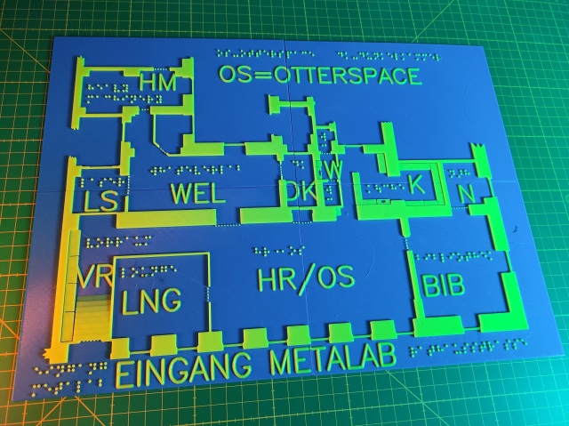 3D-Druck einer taktilen Karte vom metalab. Beschriftung in Profilschrift und Braille der Räume, Mauern, Türen, Fenster und teile der Einrichtung sind mit gelbem Kunststoff auf eine blaue Grundplatte gedruckt. Die Karte besteht aus vier rechteckigen Teilen. In Profilschrift steht auf der Karte "OS=Otterspace", "HM", "LS", "WEL", "DK", "W", "K", "N", "VR", "LNG", "HR/OS", "BIB", "Eingang metalab"
