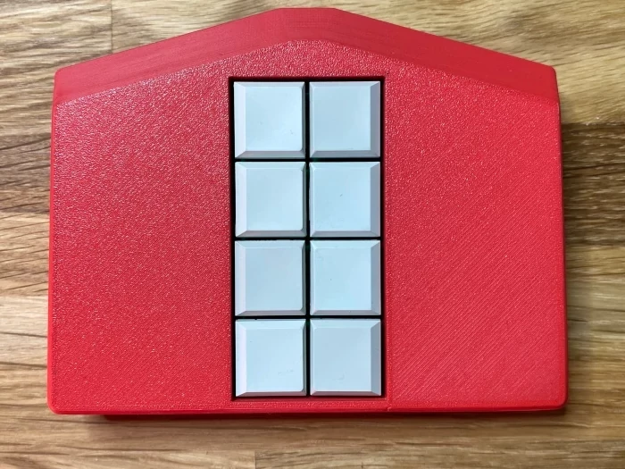 Mobile Braille-Tastatur Metabraille mit 8 Tasten. Das Gehäuse ist rot und die Tasten sind weiß. Das Gehäuse ist ungefähr fünfeckig. Ein Rechteck wird an der Breitseite durch ein Trapez erweitert. Die Form ähnelt einem Haus mit Fenstern in der Mitte.