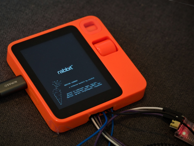 A rabbit R1 (a bright orange plastic square, with a crappy LCD panel), with jumper wires coming out of a hole in the side, showing the 'carroot' boot screen, which includes an ascii art carrot