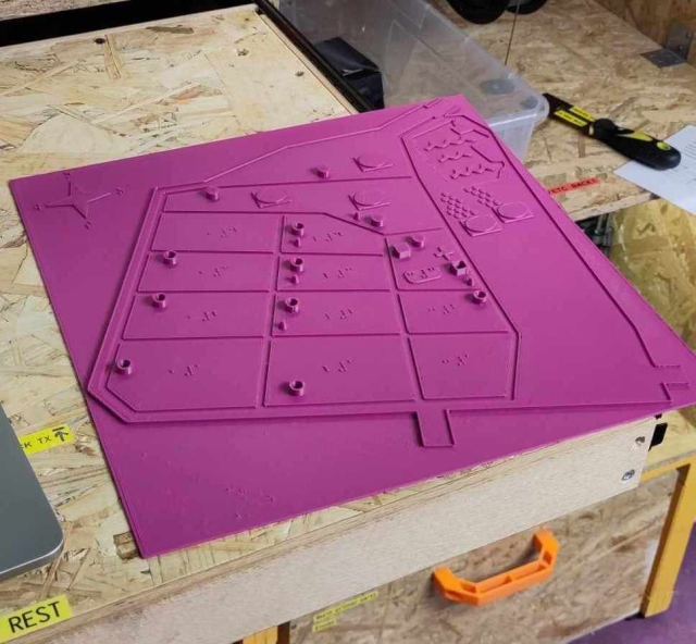 A fuchsia colored 3D print, 30cm by 30cm. There's a grid of planes with cylinders and cubes on it, also a lake area,  five stages and some braille markings