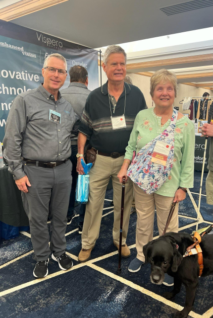 3 people standing and smiling at the Vispero booth in the ACB exhibit hall. Left to right; Bill Kilroy, Mark Lear, Kati Lear with her guide dog.