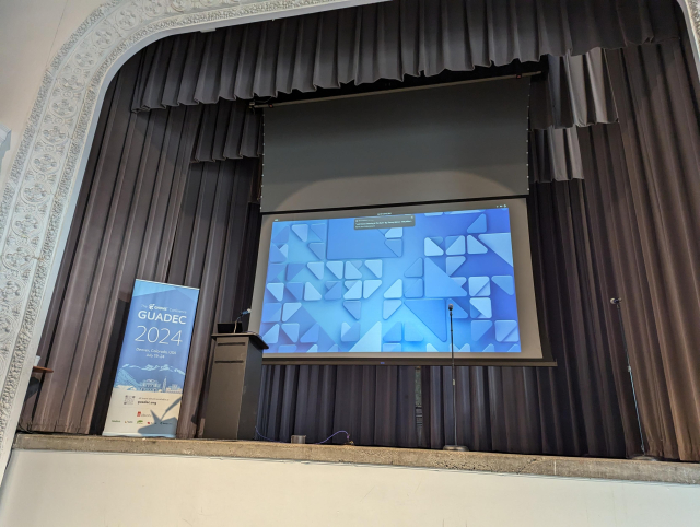A small stage, recessed into the wall and bordered by ornate crown molding. A banner on stage has the logo and text for GUADEC 2024, and stands by an empty podium. A screen is rolled down behind the podium, and has a matching blue geometric background to the banner.