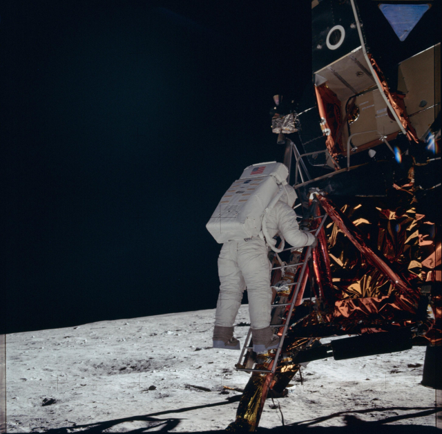 Color photo of an astronaut descending the ladder from the LM. He is wearing a puffy white suit with a large, rectangular backpack. Parts of the LM are covered in a copper-colored foil.
