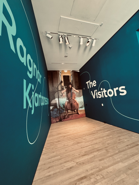 Two blue walls lead to a floor to ceiling image of a young woman with brown hair playing a cello. The left wall says Ragnar Kjartansson and the right one says The Visitors. 