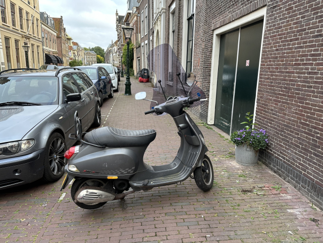 Picture of a grey Vespa blocking the sidewalk in between parked cars and buildings.