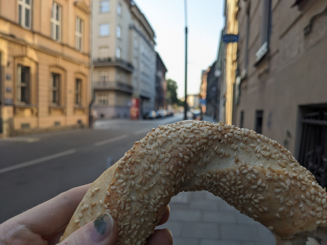 A sesame-covered, ring-shaped bread, held in front of a street with beige houses.