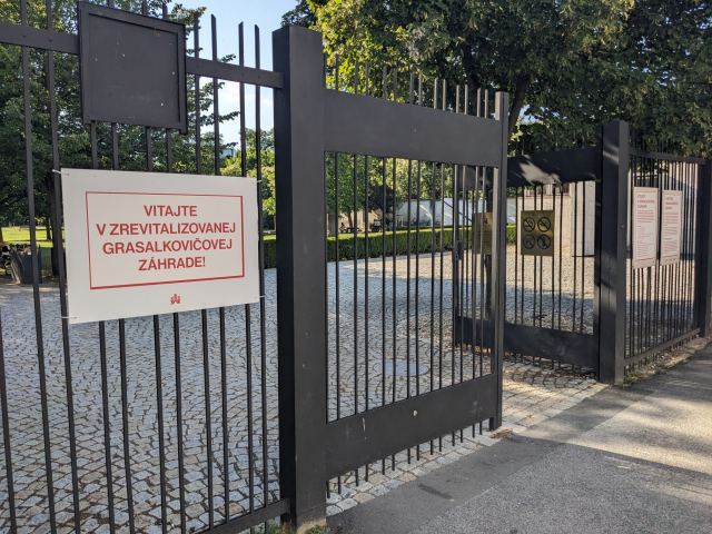 A sign on a fence says "VITAJTE V ZREVITALIZOVANEJ GRASALKOVIČOVEJ ZÁHRADE!"