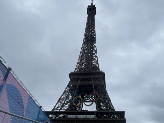 the Eiffel Tower in Paris, France. The photo is taken from a low angle, looking straight up at the tower. It is a cloudy day with a gray sky as the background. The Eiffel Tower itself is a tall, iron lattice structure, and prominent in the image are the five interlocking Olympic Rings hanging below one of its lower levels. The Olympic Rings are colored blue, yellow, black, green, and red. 