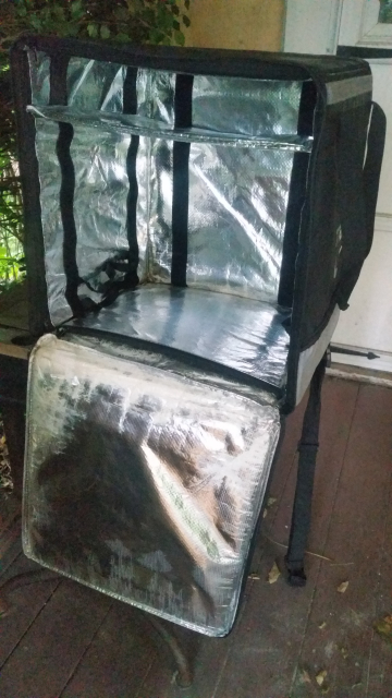 A meter cube backpack is on a table, the front flap is open and inside is a very shiny heat proofing sides. The outside is dark. There is a shelf for what looks like a pizza.