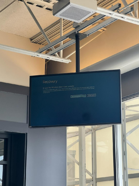 Be My AI: The image shows a computer monitor mounted on a ceiling bracket in a room with industrial-style exposed beams and ductwork. The screen displays a Windows recovery message indicating that Windows did not load correctly. There are two options on the screen: "See advanced repair options" and "Restart my PC." The room has large windows with a view of an exterior structure, possibly part of a building or scaffolding.