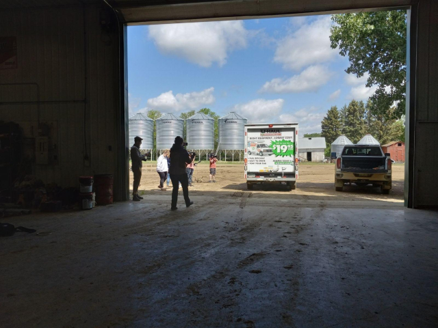Several reporters out in a farm yard with rows of grain bins and trees, filming as the Uhaul truck with space junk inside drives away