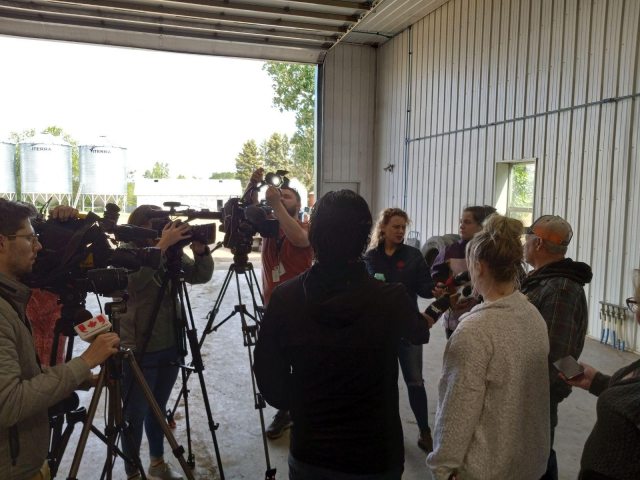 A tight cluster of TV cameras and mic-holding reporters all focused on the farmer.  Everyone is standing in a large open farm shop building.