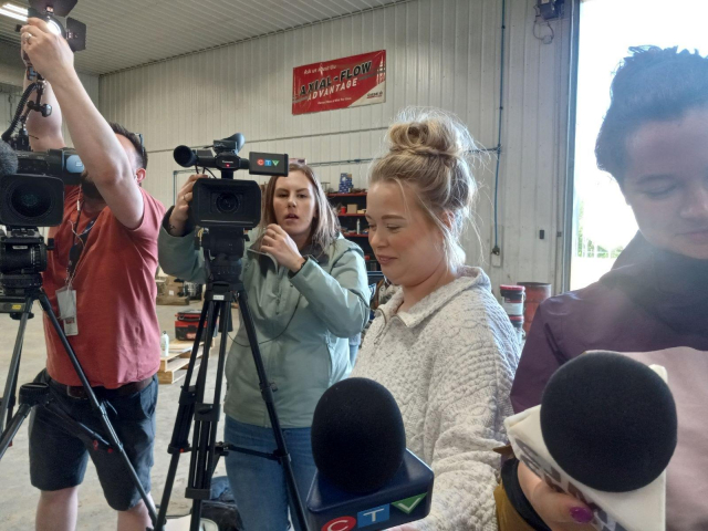 2 journalists setting up TV cameras and lights facing toward me, and 2 more pointing mics into my face. We're all inside a large metal farm shop building.