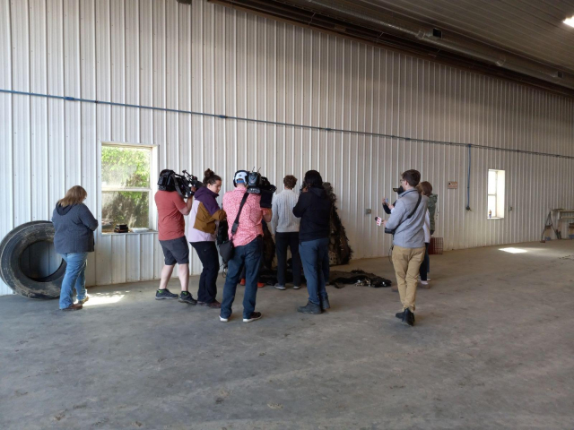 A crowd of journalists, most carrying large TV cameras, swarmed around 2 engineers looking at the pile of space junk, inside the big metal farm shop building.
