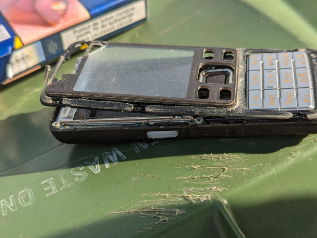 A broken Nokia mobile phone laying on a green surface. The case is busted, the front bezel is pushed up, the joystick is smashed. 
It probably still works. 