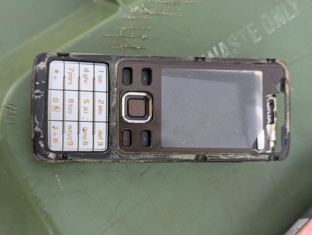 A broken Nokia mobile phone. A black rectangle with a small clear rectangle (the screen) below that a series of holes where buttons would be (removed). Below that is the dialing pad, a 4x3 array of white buttons with red text. 