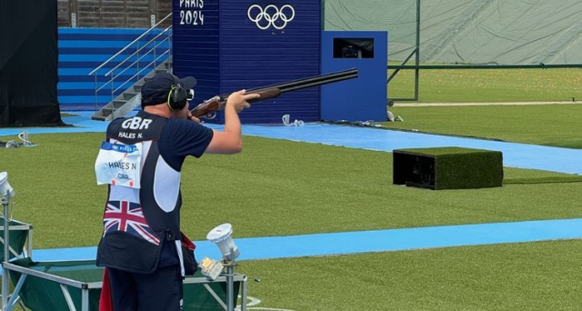 nathan Hales depicted here at the paris Olympics 2024, wearing a blue shirt, a cap and hearing protection whilst holding his gun