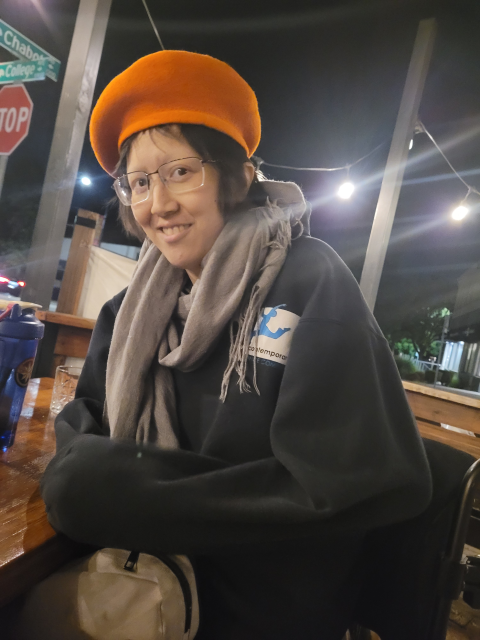 Mel Chua smiling at camera while wearing an orange hat, seated at an outdoor restaurant.