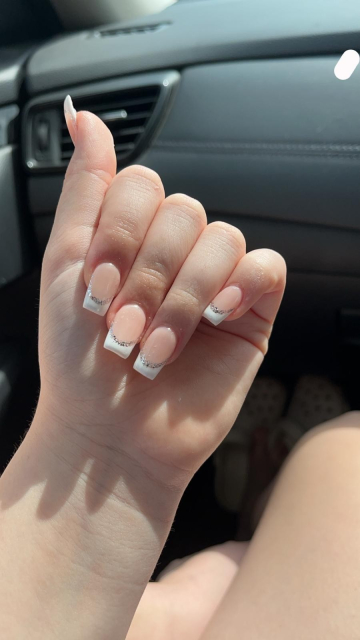 a close-up of a hand with a manicured set of nails. The nails are short to medium in length and square shaped. The base color of the nails is a natural-looking nude tone, while the tips feature a classic French manicure style with white tips. Additionally, there is a subtle silver glitter line that separates the nude base from the white tip, adding a touch of sparkle. The hand is held up against the interior of a car, specifically near the dashboard and a vent on a sunny day.