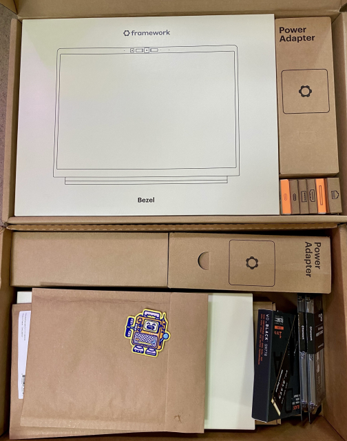 An open box containing various packaged electronics items including a "framework" laptop bezel, power adapter, cables, and a few accessories. There is also a brown paper envelope with a colorful robot sticker on it.