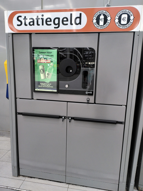 A grey rectangular machine with various locked doors. In the middle is a black panel with an oblong shape gloss panel, a round black panel. A silver knob. And a screen.