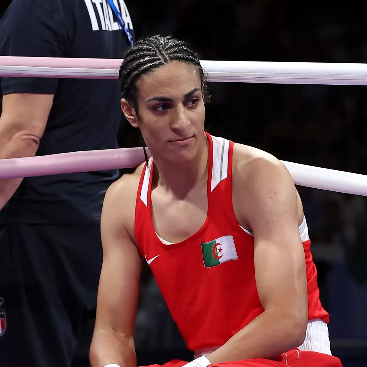 Close shot of Imane Khelif, sitting down, wearing a red uniform with the Nike symbol and the Algerian flag, and looking sideways.