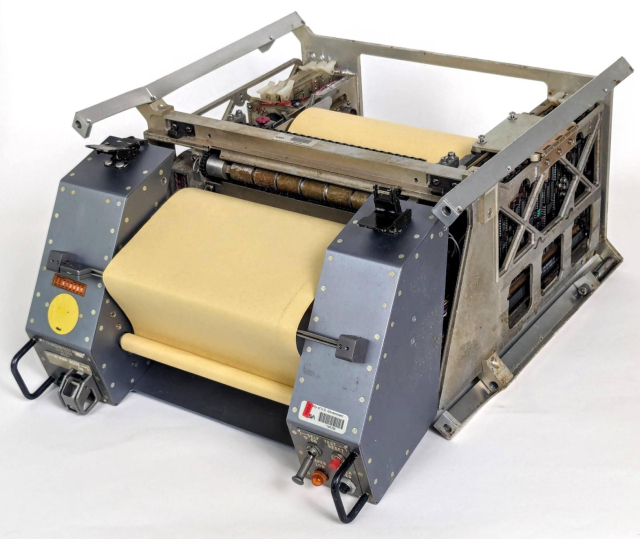 The teleprinter is a large device, a bit smaller than a microwave, with a roll of yellow paper running through it. The teleprinter has rails for mounting in a locker. It has some controls and switches on the front, which is grayish metal.