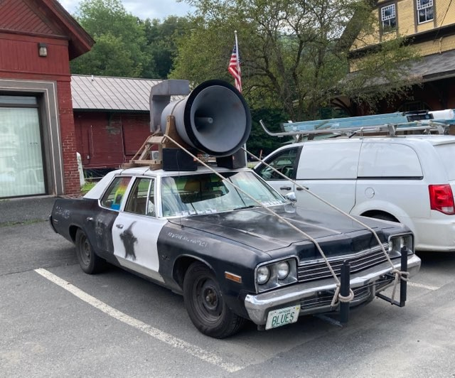 A car parked in a Vermont village which is a black and white car looking like a police cruiser with a HUGE speaker roped to the top of it, in the style of the movie,