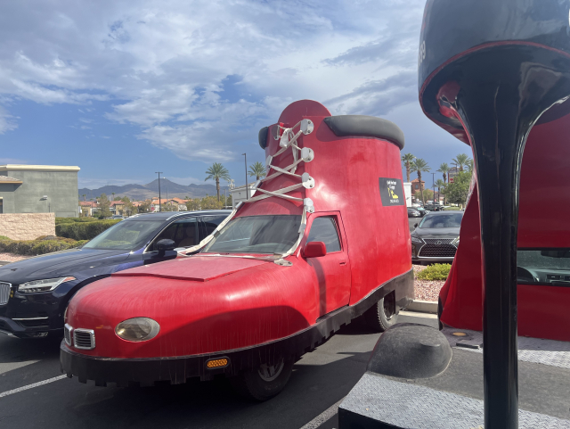 A red car modified to resemble a large shoe, parked in a lot with other vehicles under a partly cloudy sky.