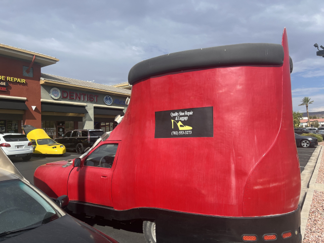 A car shaped like a large red boot parked in front of a strip mall with a dentist's office and other businesses.