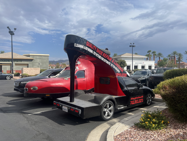 A unique, car-shaped advertisement vehicle for a shoe repair and upholstery business is parked in a commercial parking lot. The vehicle is shaped like a high-heeled shoe and has contact details on its side. Other cars and stores are visible in the background