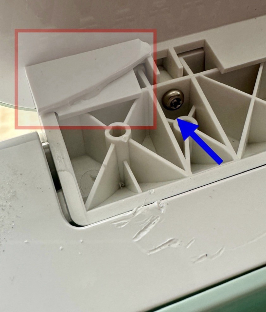 Close-up showing a broken piece of white plastic near a screw inside an appliance, with a blue arrow pointing to the screw.