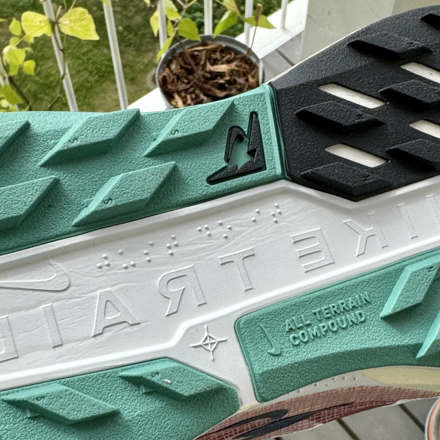 The sole of a green and white running shoe with some braille visible. Other marks include the words “Nike Trail”, Nike’s “swoosh” logo and the words “all terrain compound”. 