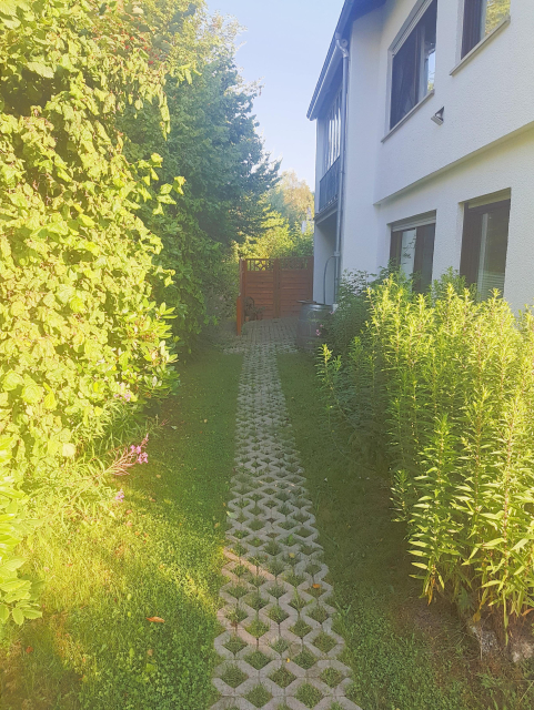 Weg aus Rasengittersteinen. Links sind sehr hohe Bäume und Büsche, rechts Stauden und ein Haus.
Der Weg endet vor einem Eingang.