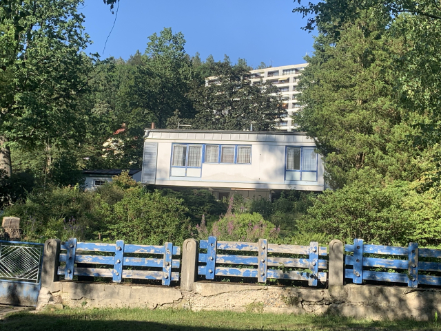 A house with a white and blue exterior situated on a hillside, surrounded by greenery and trees. In the foreground, there is a weathered blue and stone fence.