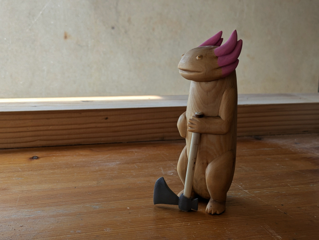 A woodcarved standing axolotl carrying an ax. The ax is in its left hand, iron of the tool to the ground.